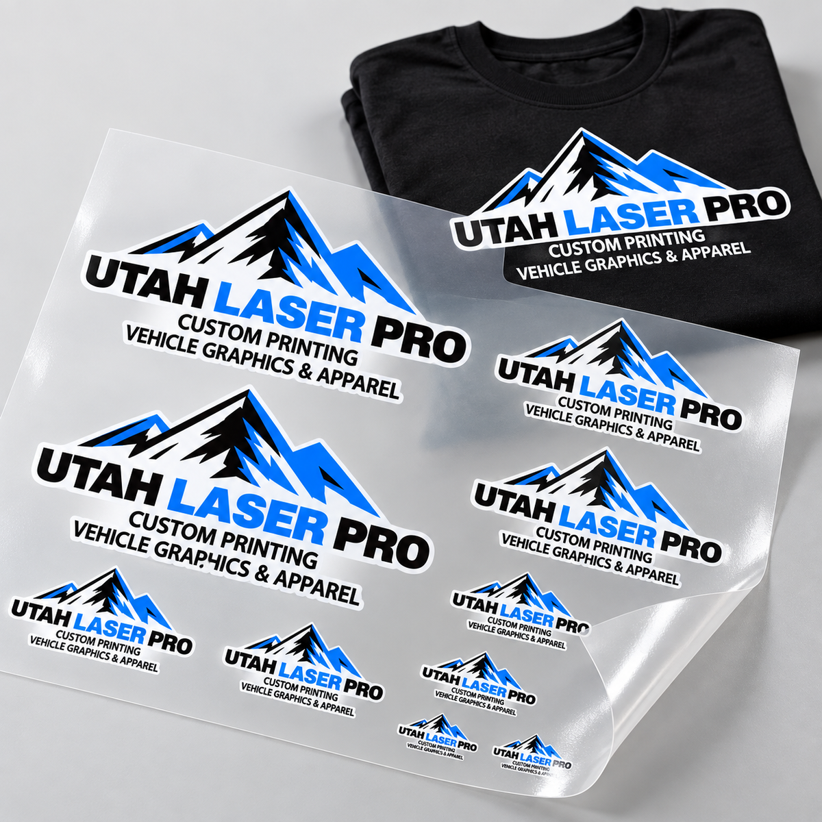 Mockup of Utah Laser Pro logo transfers in multiple sizes on a DTF gang sheet beside a matching black T-shirt.